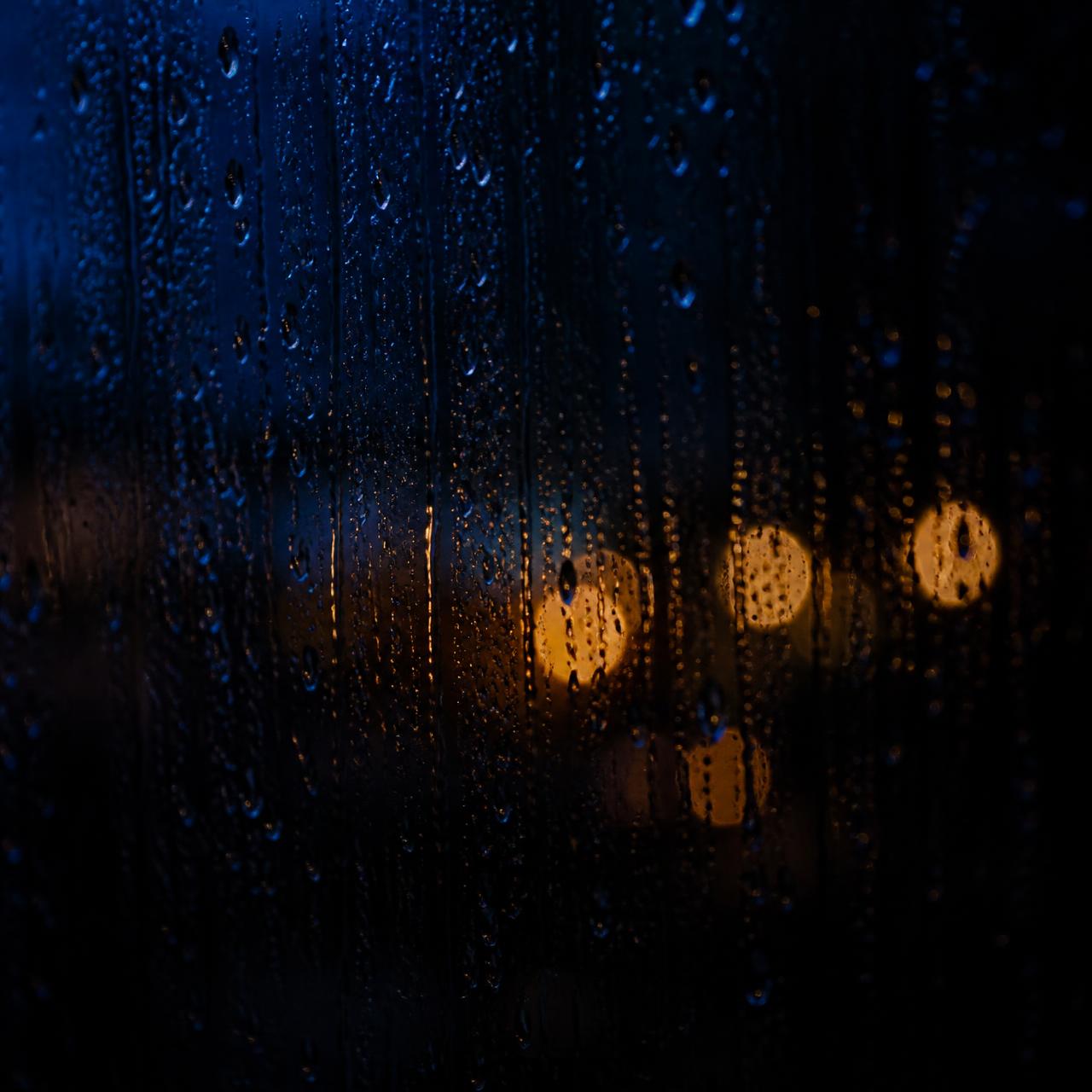 Rain running down a dark window with soft blurred amber lights behind the glass