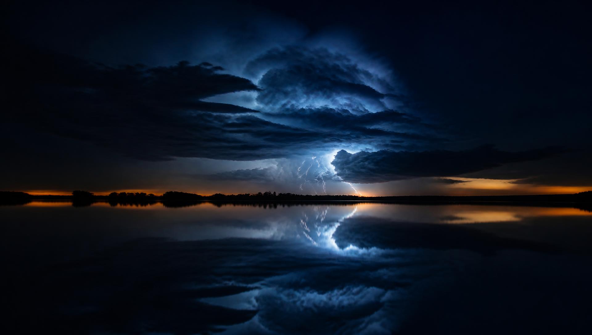 A distant thunderstorm lighting the clouds above a still lake, tree silhouettes on the far shore and the storm mirrored in the water